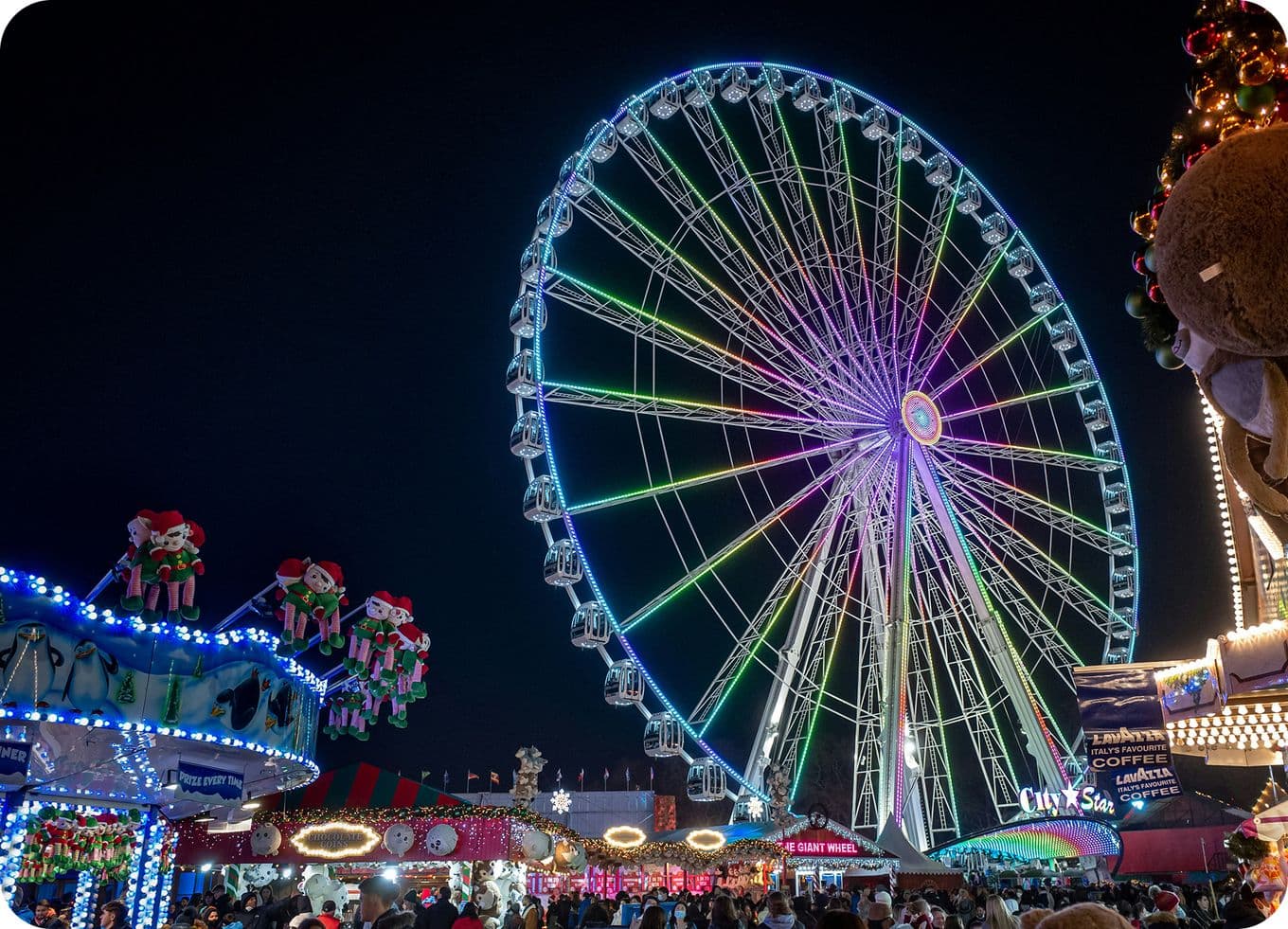 Giant illuminated Ferris wheel glowing in rainbow lights over a bustling nighttime fair with rides, booths, and crowds.