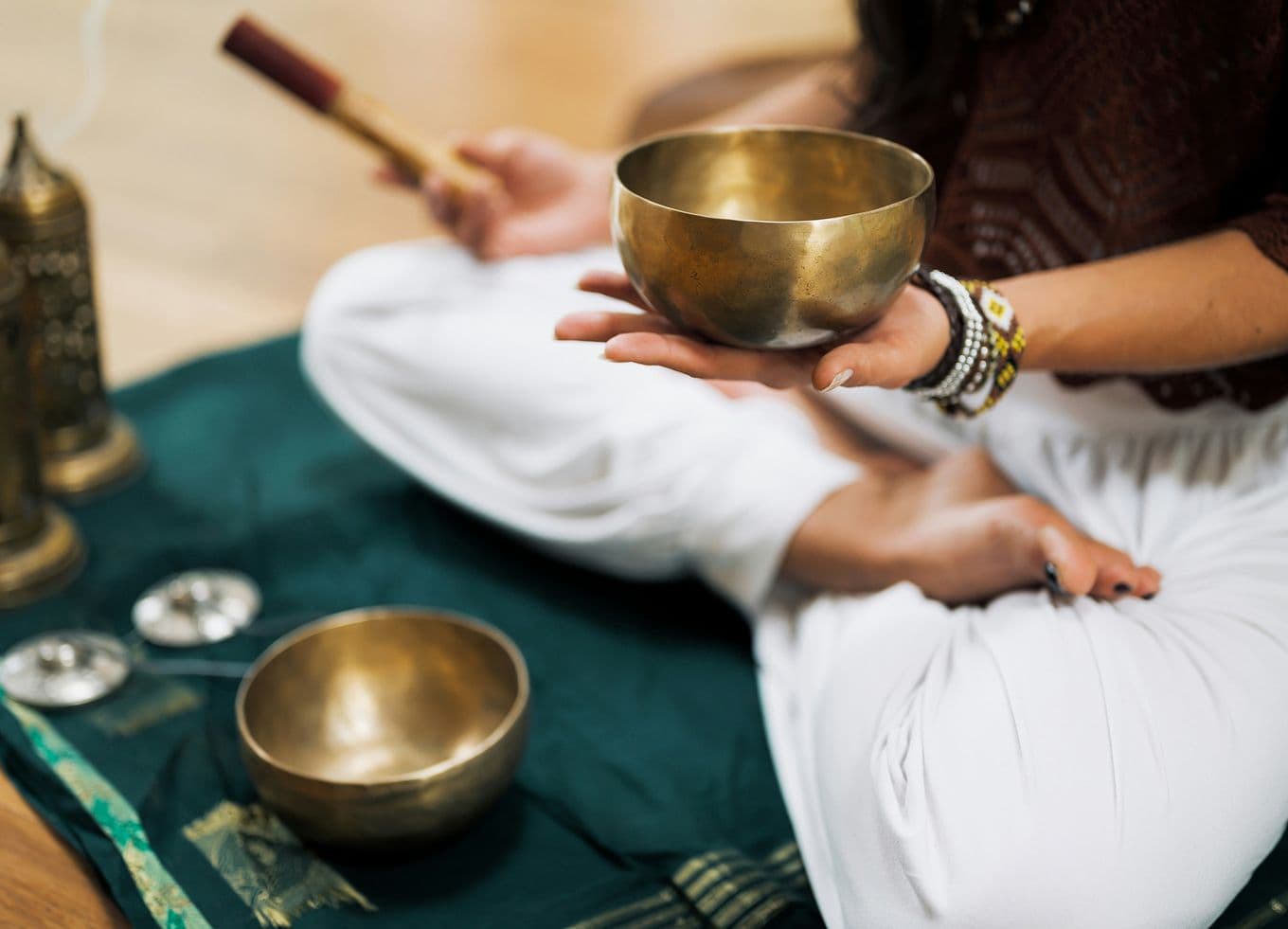 Person seated cross-legged holds a brass singing bowl and mallet, surrounded by additional bowls and ritual items on a green cloth.