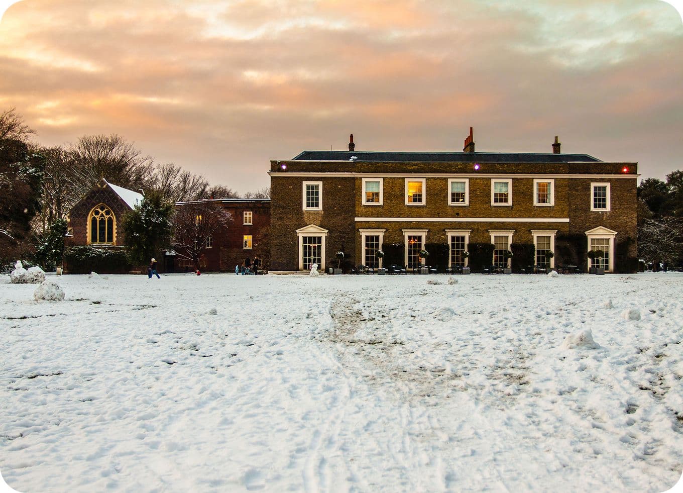 Georgian brick house with lit windows beside a small chapel, snow-covered lawn and a few people under a pink sunset sky.