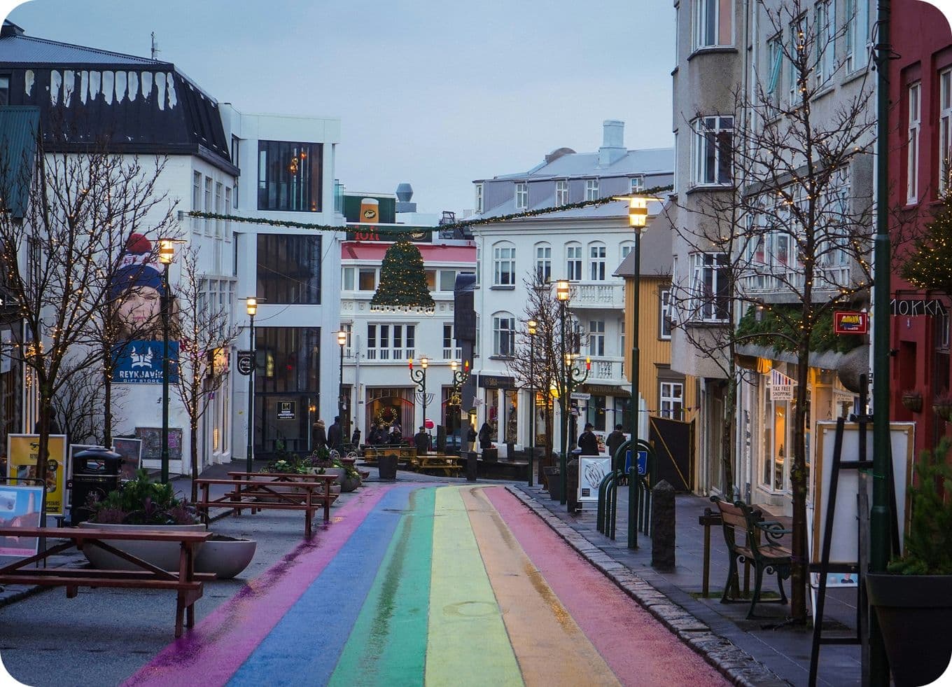 A street with a rainbow-painted path, lined with shops and bare trees, under a cloudy sky. Warm streetlights illuminate the scene.