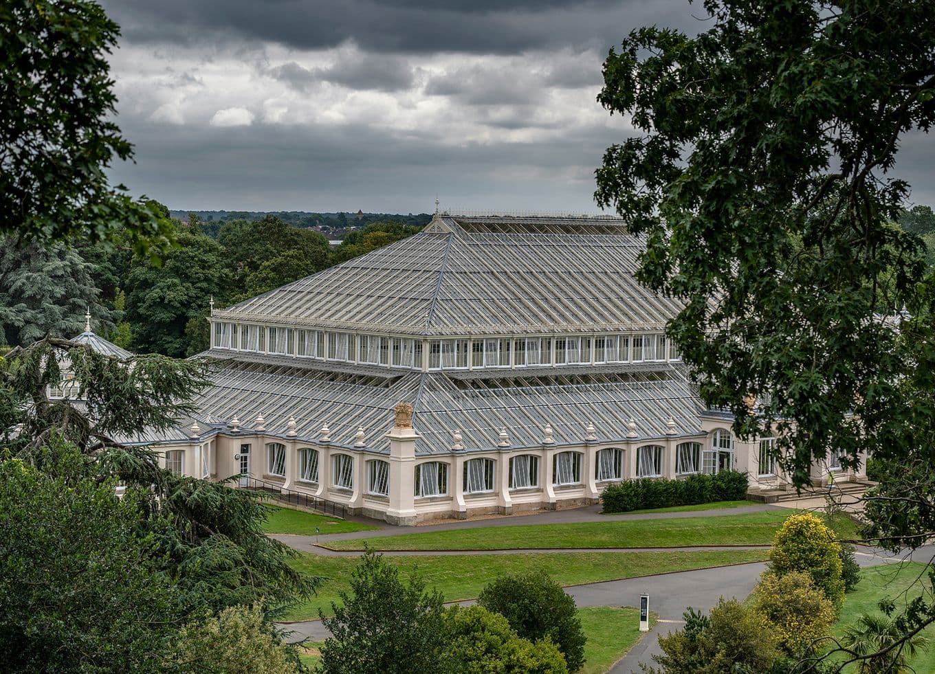 A large glass greenhouse with a metal frame and a pointed roof, surrounded by lush greenery under a cloudy sky.
