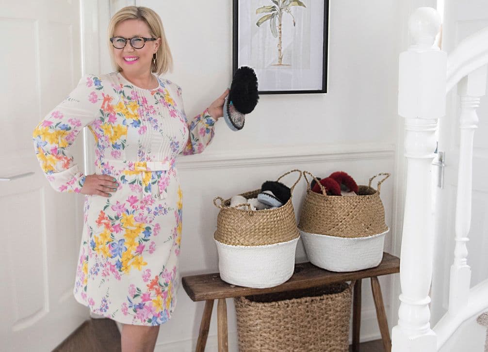 Smiling woman wearing glasses and a floral dress holding a black shoe beside woven baskets of footwear on a hallway bench.