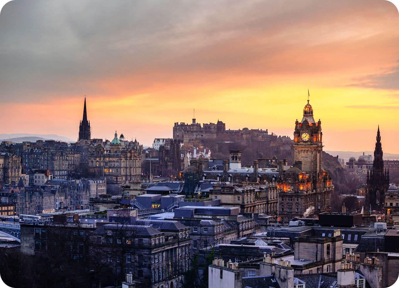 Edinburgh skyline at sunset, featuring historic buildings, a clock tower, and a distant castle under a colorful sky.