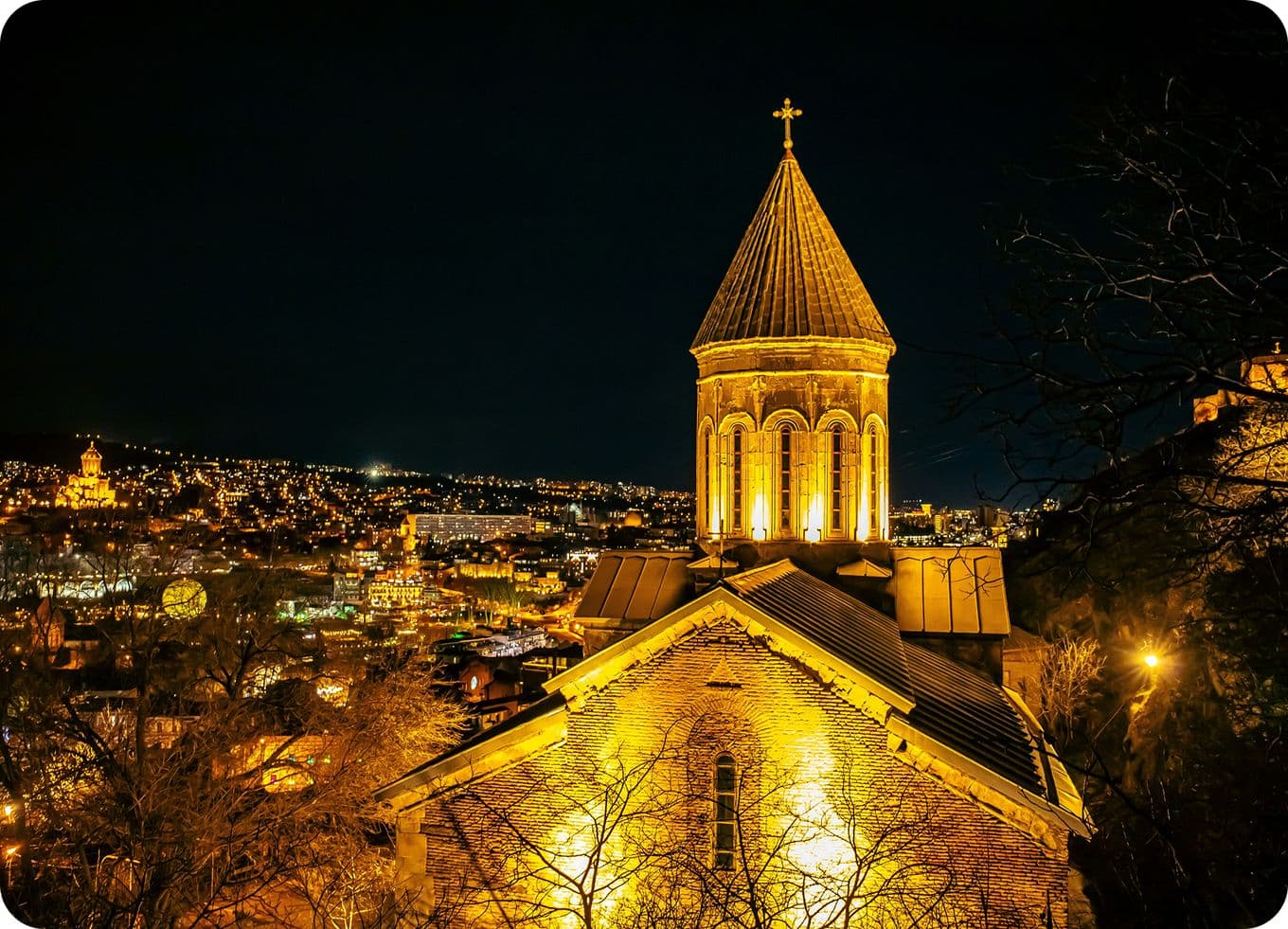 Illuminated church with a tall spire at night, overlooking a cityscape with glowing lights and a dark sky.