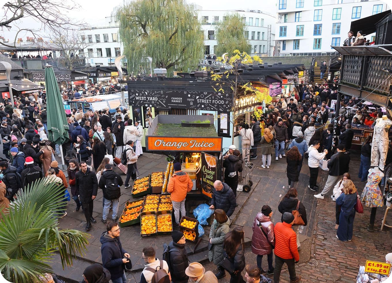 Crowded outdoor market with an orange juice stand, stacks of oranges and shoppers milling among stalls and surrounding buildings.