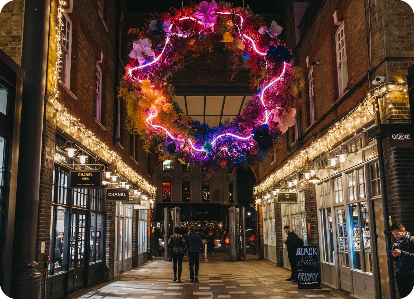 Nighttime shopping arcade lit with string lights, a large neon-lit floral wreath suspended above shoppers walking between boutique storefronts.