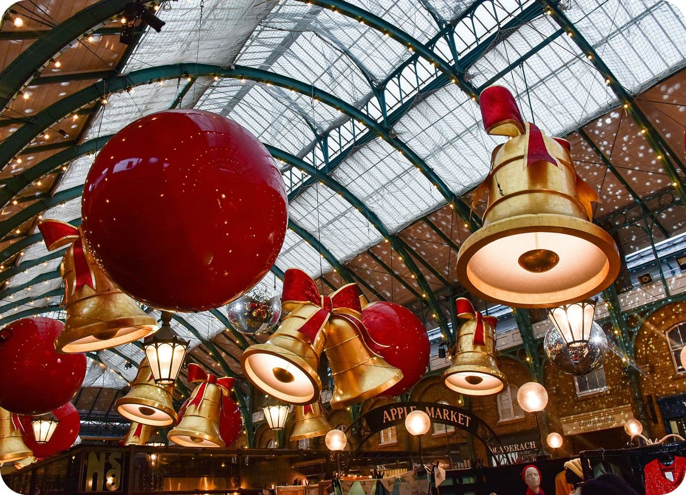 Covered market interior decorated for Christmas with oversized gold bells and giant red baubles under a glass arched roof.