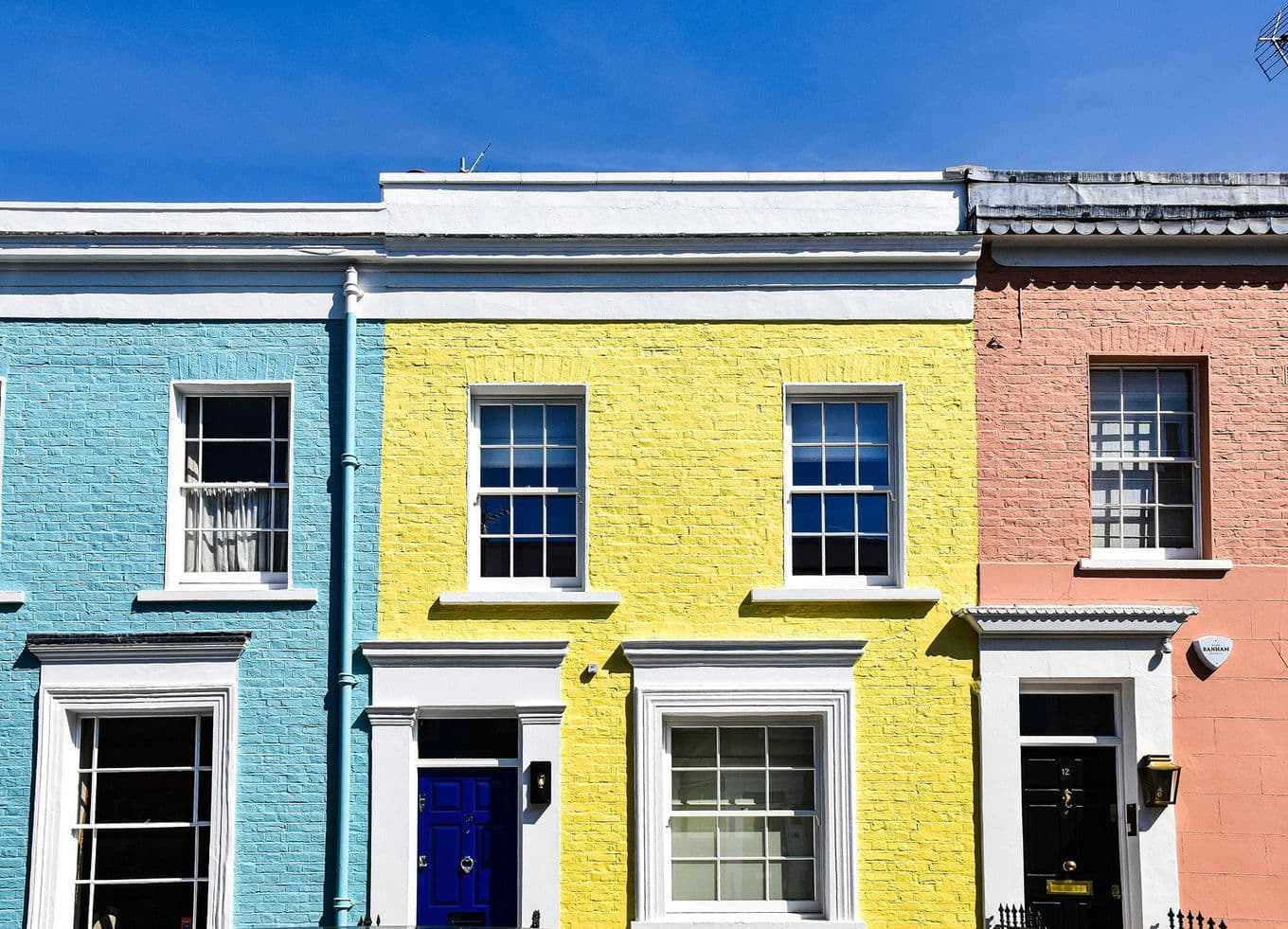 Three colorful row houses with blue, yellow, and pink facades under a clear blue sky. Each house has white-framed windows and doors.