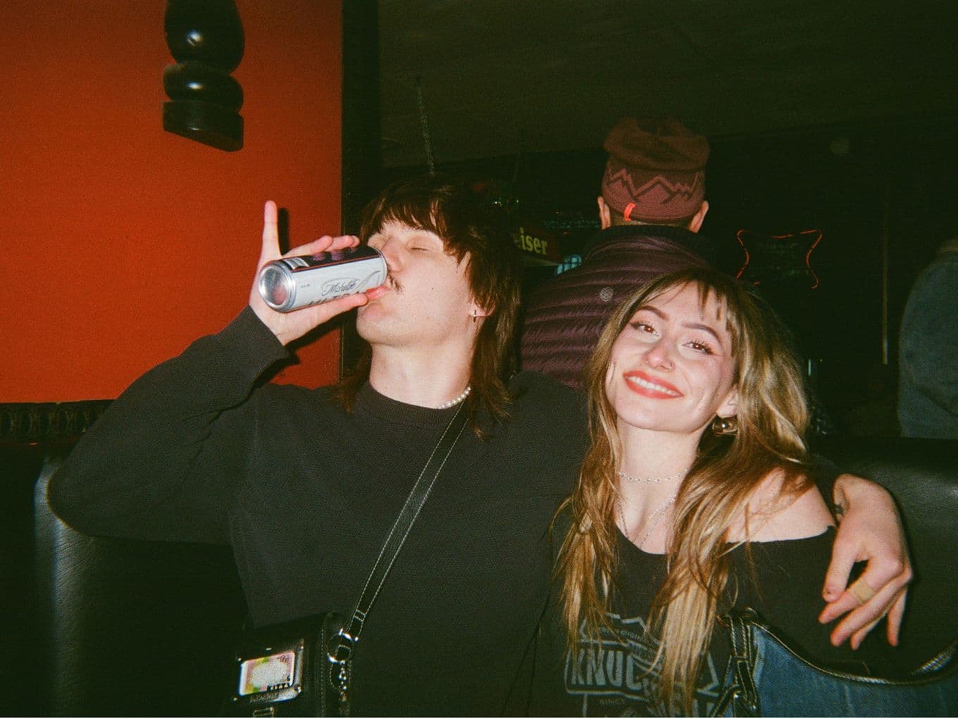 Two people sitting in a dimly lit setting; one is drinking from a can, and the other is smiling widely at the camera.