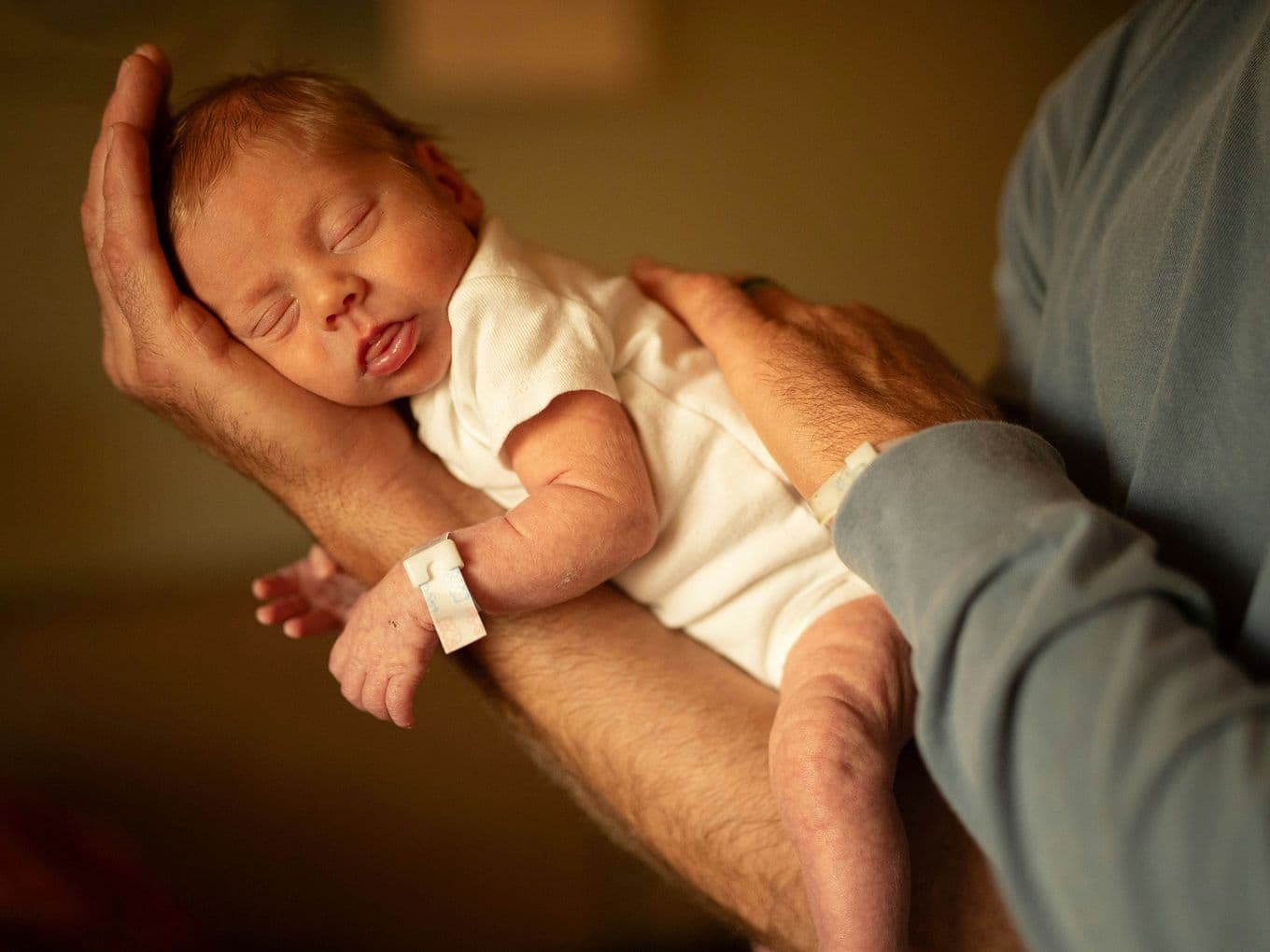 Newborn baby wearing a hospital wristband, peacefully asleep on a person's arm in a cozy, soft light setting.