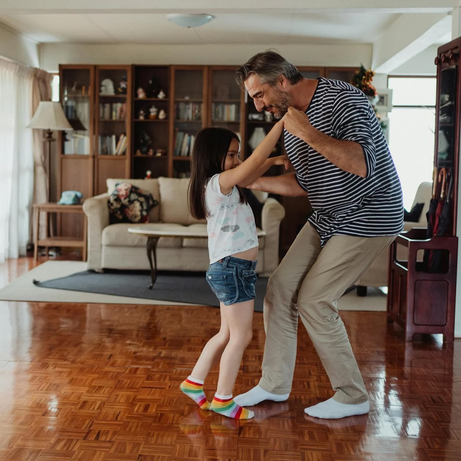 A man and his young daughter dance joyfully in a living room with wooden floors