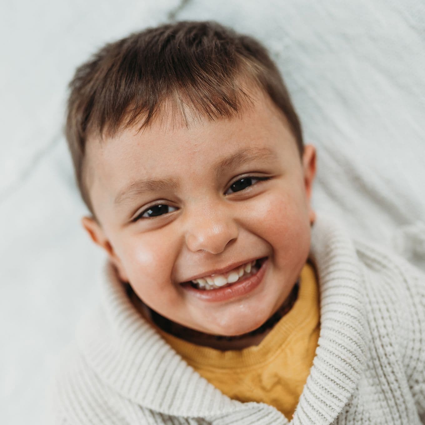 Preschool-age boy smiles looking up at the camera while laying on a bed