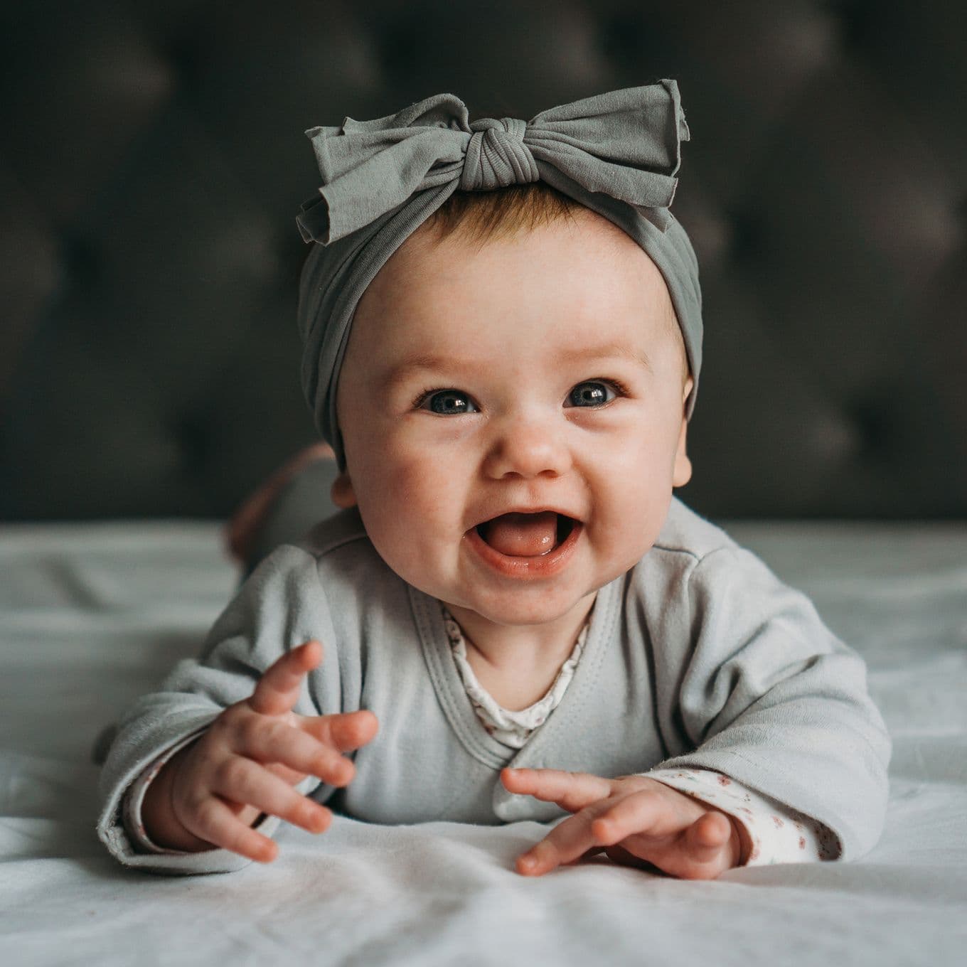 A young baby girl with a bow in her hair smiling at the camera while laying on her belly during a baby photoshoot