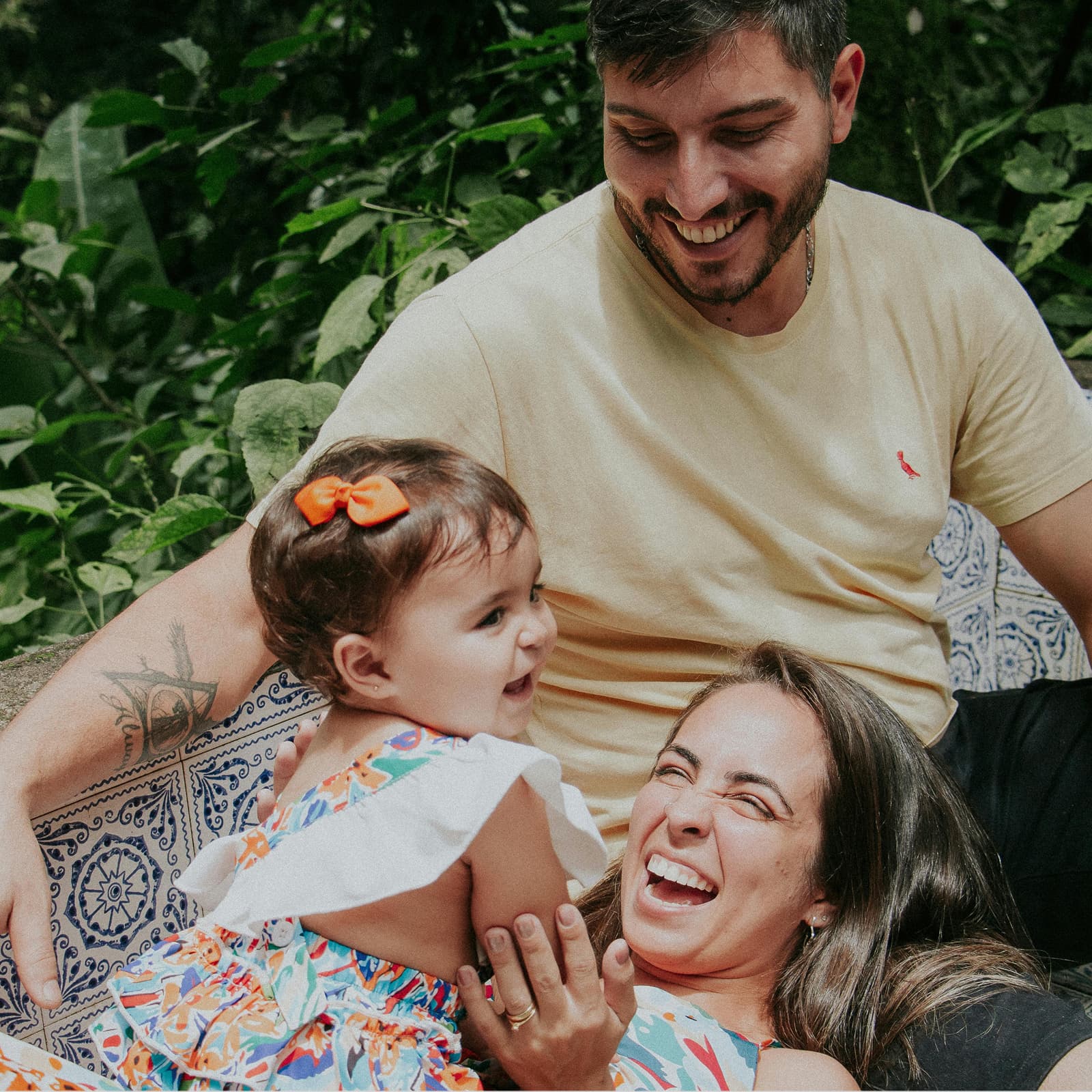 A man, woman and toddler with a bow enjoy a joyful moment together outdoors, surrounded by greenery.