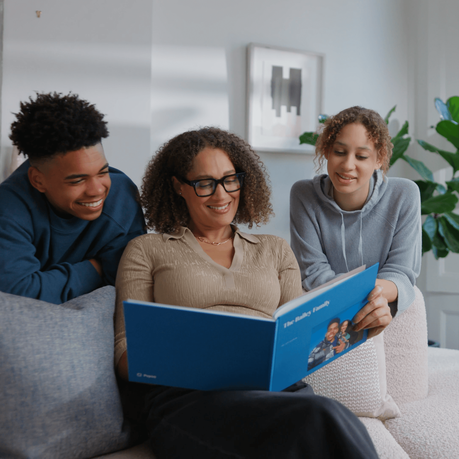 A woman and two teenagers smile while looking at a large blue Popsa Photo Book on a couch in a cosy living room