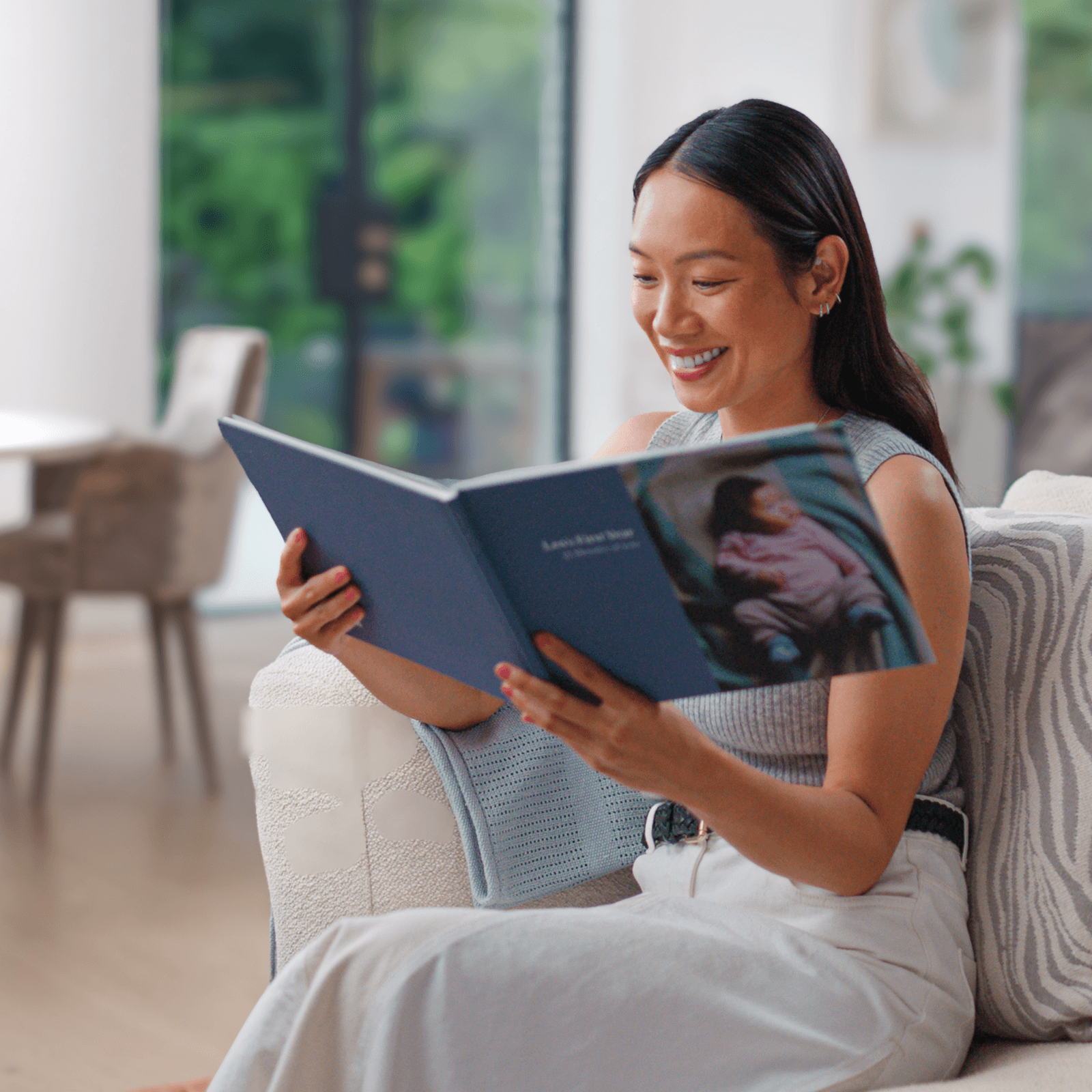 Smiling woman sitting on a couch, reading a large photo book in a bright, modern living room