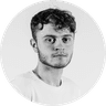 Young man with short curly hair and a beard, wearing a white t-shirt, against a neutral background, looking directly at the camera.