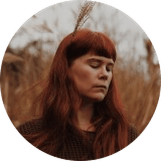 Red-haired woman with eyes closed in a dry grass field, a single reed tucked in her hair.