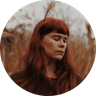 Red-haired woman with eyes closed in a dry grass field, a single reed tucked in her hair.