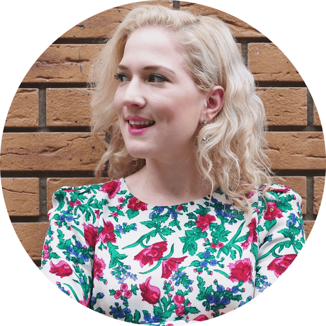 Young woman with wavy blonde hair smiling, wearing a white floral dress, looking left against a brick wall.