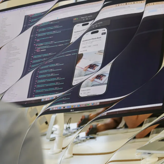 Distorted view of a person working on a monitor showing code and a mobile app preview, with a laptop displaying app listings nearby.