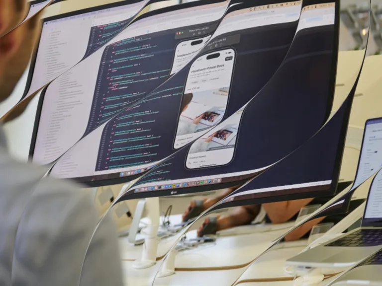 Distorted view of a person working on a monitor showing code and a mobile app preview, with a laptop displaying app listings nearby.