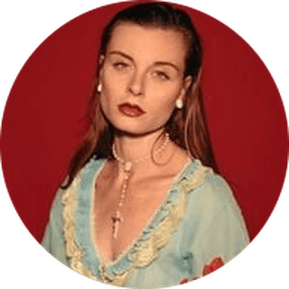 Woman with long hair, wearing a light blue dress with lace trim and a necklace, stands against a red background.
