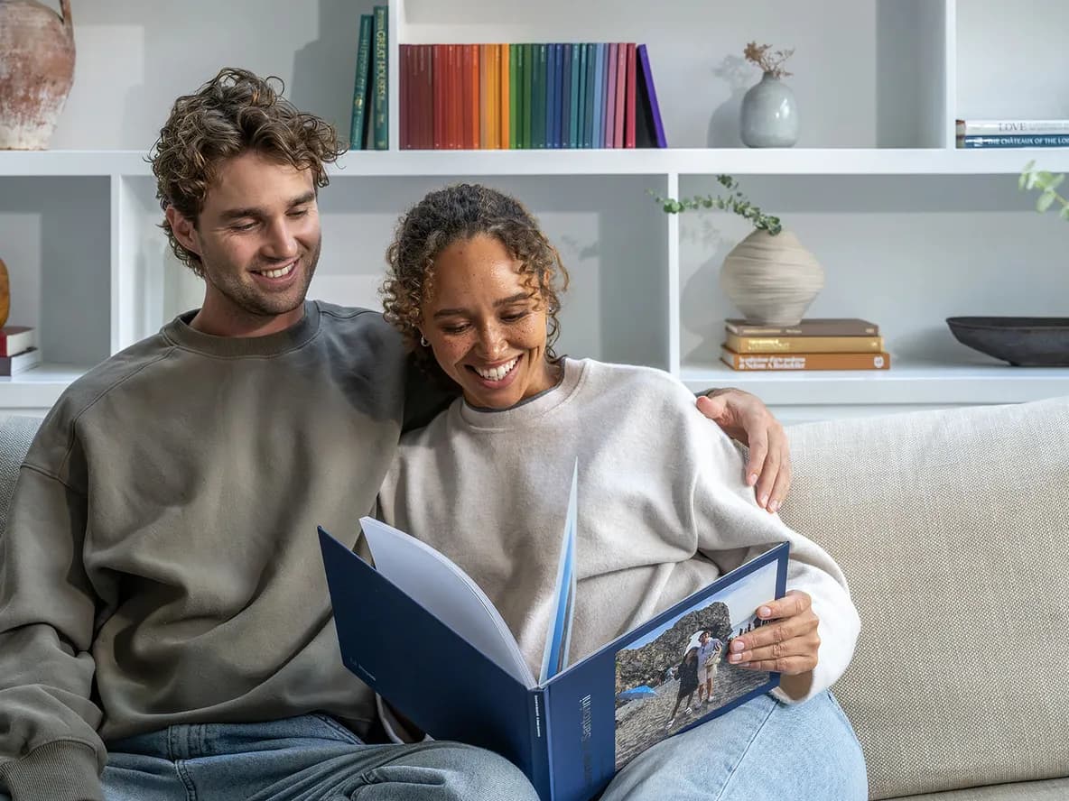 A smiling couple on a sofa looks at a Popsa photo album, open to a picture of a rocky beach. White shelves with books and decor are in the background.