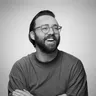 A bearded man in yellow glasses and an olive green shirt laughs with arms crossed against a grey background.