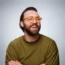 A bearded man in yellow glasses and an olive green shirt laughs with arms crossed against a grey background.