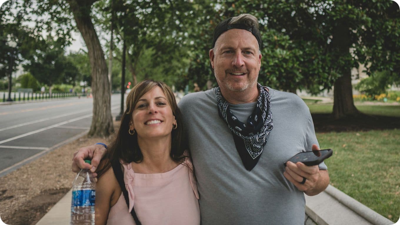 A smiling couple stands outdoors; the man holds a phone and wears a bandana, while the woman holds a water bottle. Trees line the background.
