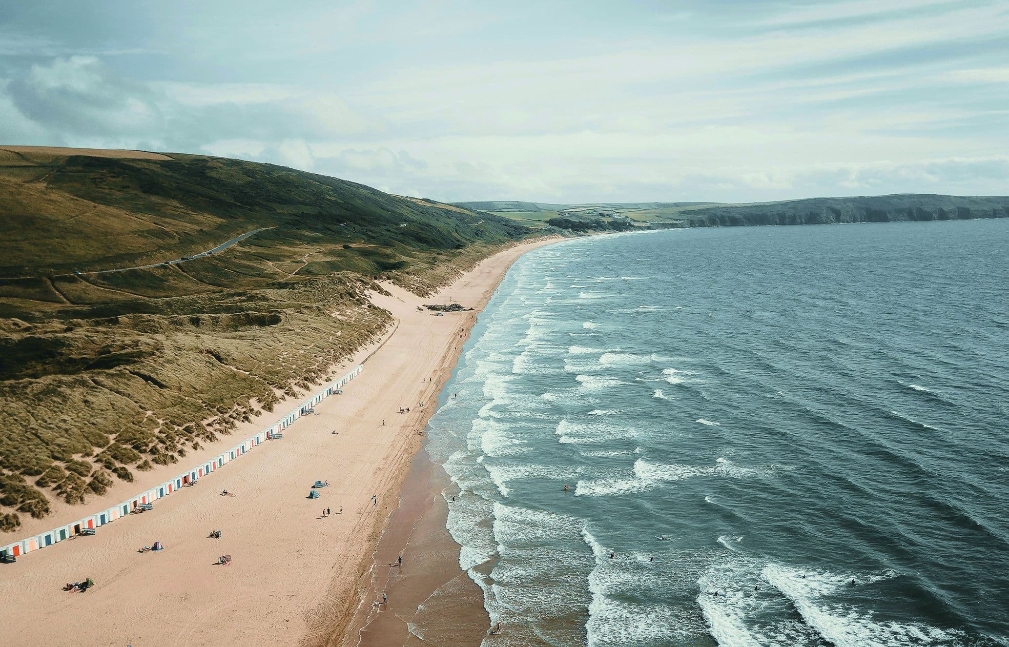 Aerial view of a sandy beach lined with colorful beach huts, gentle waves meeting the shore, and grassy hills in the background under a partly cloudy sky.