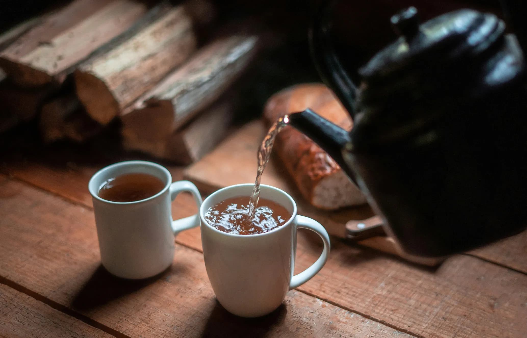Two white mugs on a wooden table, one being filled with tea from a black kettle, with stacked logs in the background.