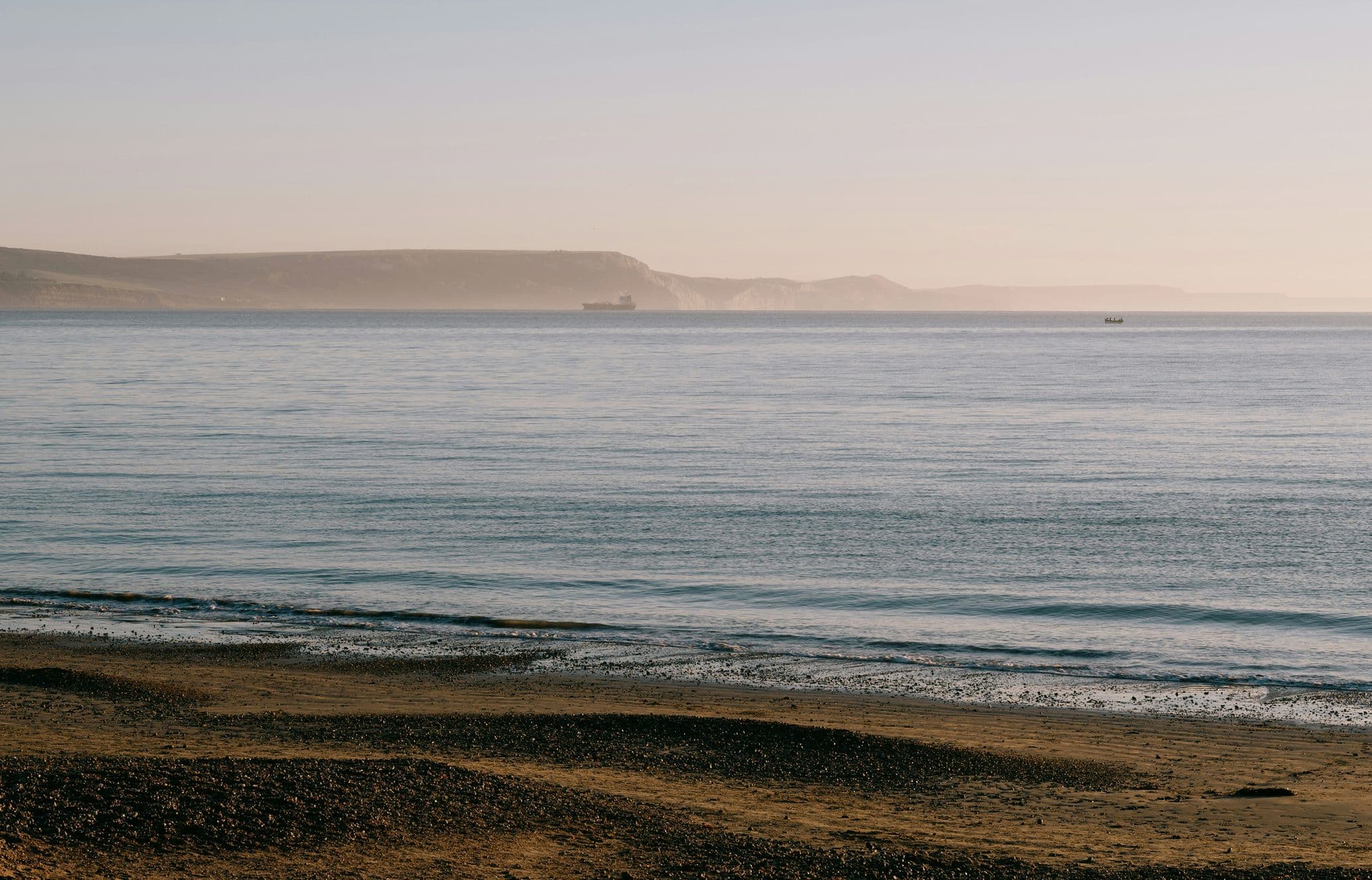 A tranquil beach scene with a sandy shore, calm ocean waves, and distant cliffs under a clear sky.