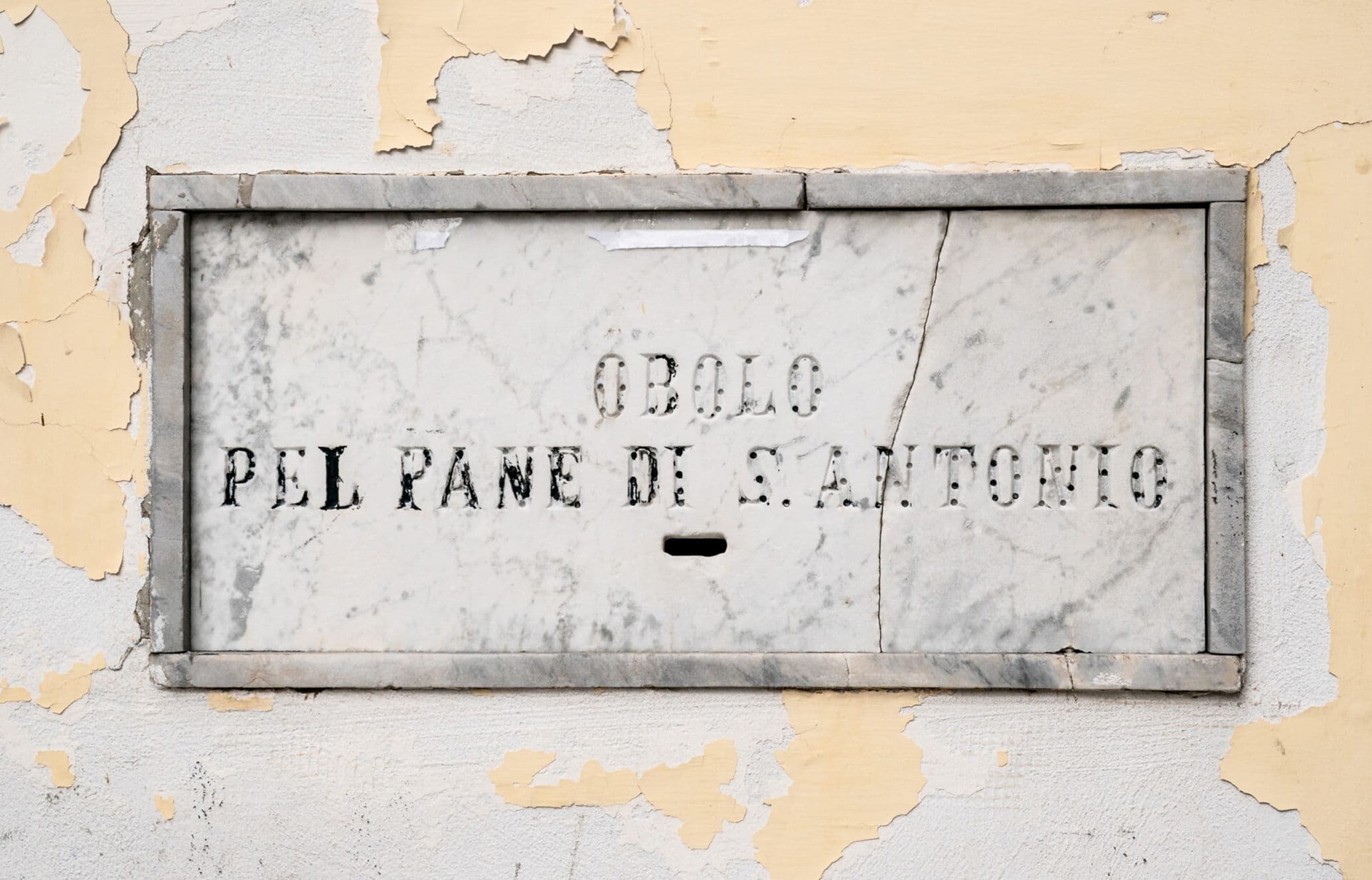 A weathered marble donation box on a cracked wall, with Italian text reading "Obolo pel pane di S. Antonio.”