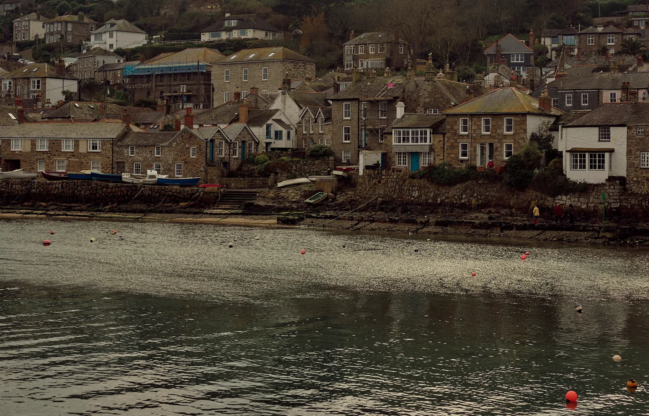 A small coastal village with stone houses, boats docked along the shoreline, and a calm water body with buoys. Sparse vegetation on the hills.