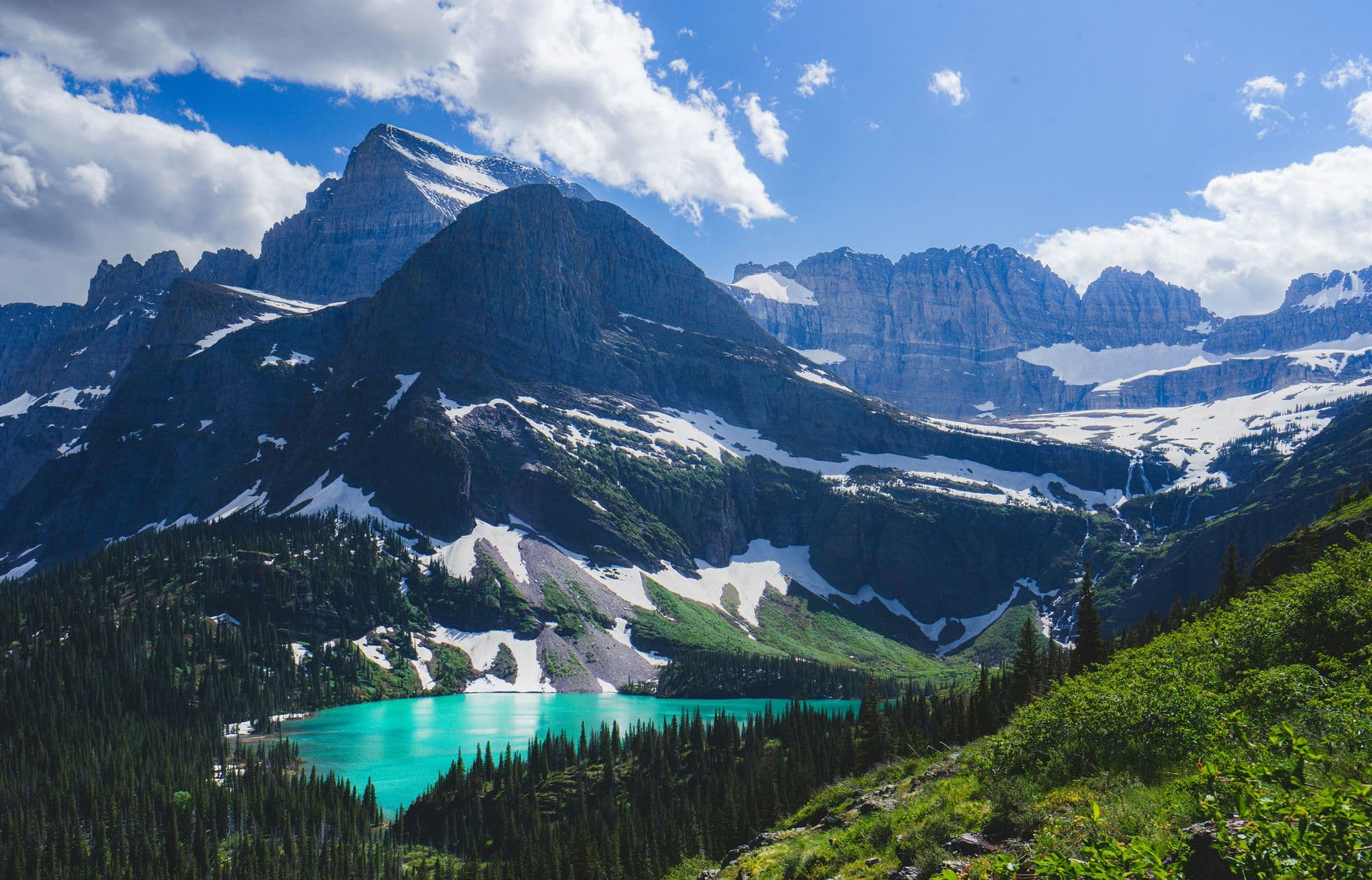 Mountain landscape with a turquoise lake, snow-dusted peaks, and lush green valley under a blue sky with scattered clouds.