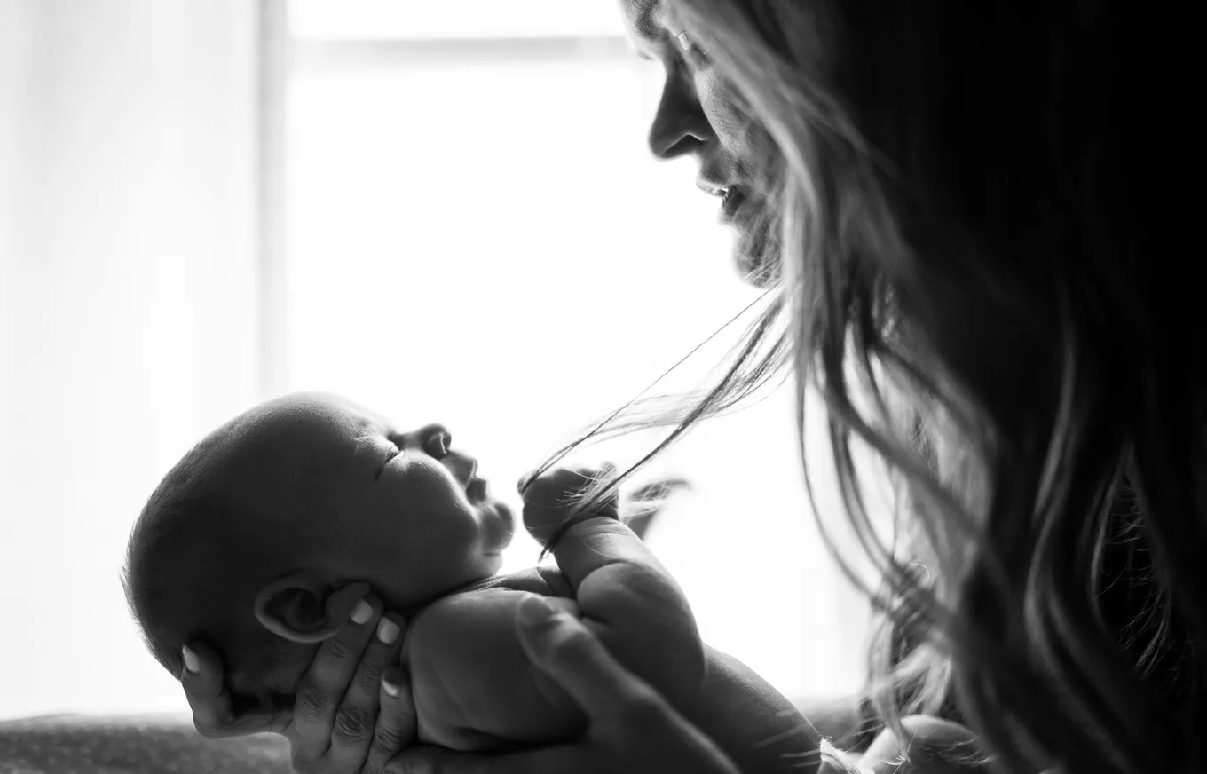Woman holding a newborn baby, smiling in close interaction, in a warmly lit room.
