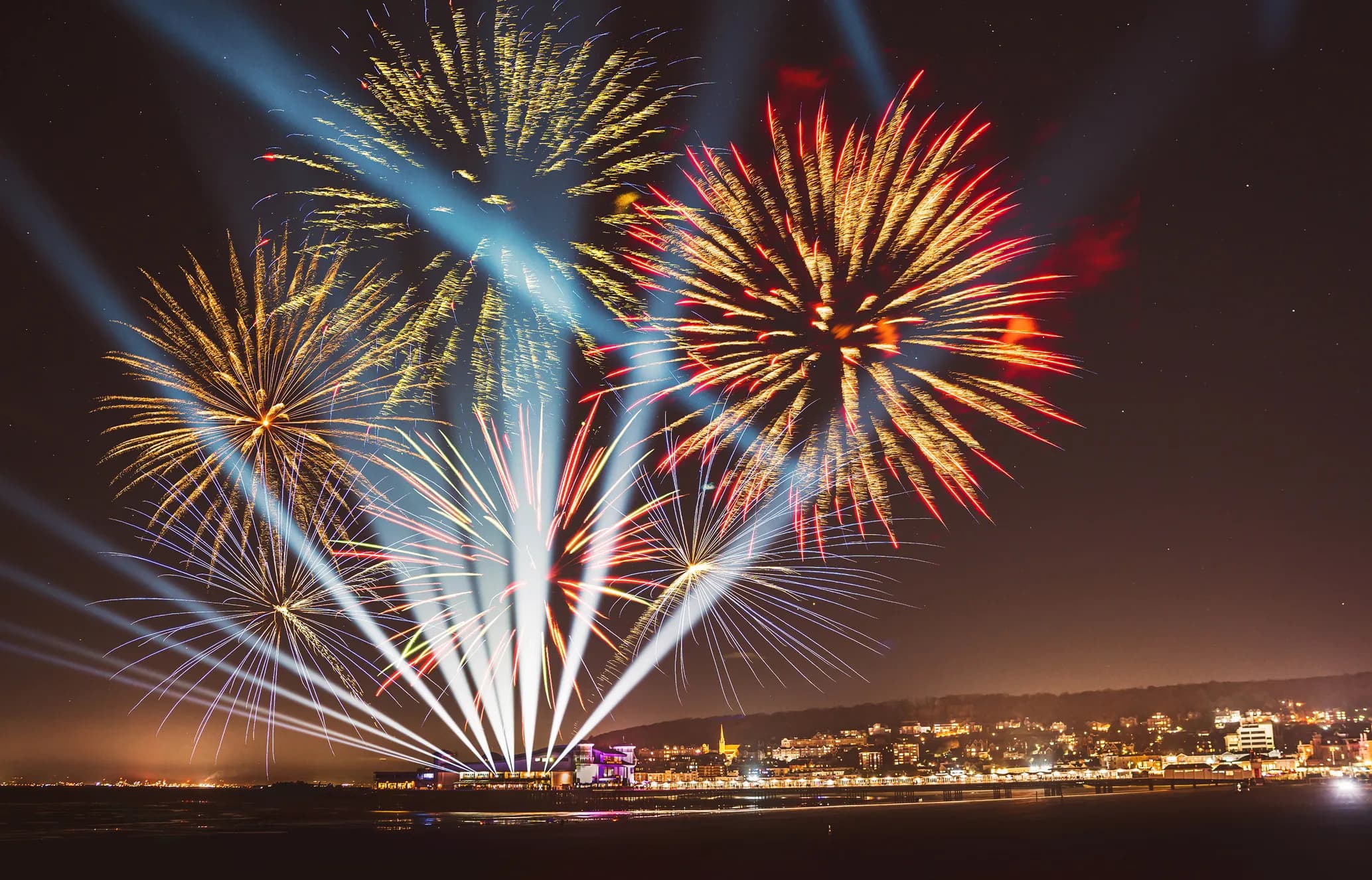 Colorful fireworks illuminate the night sky above a town, with bright beams of light and reflections on the water.