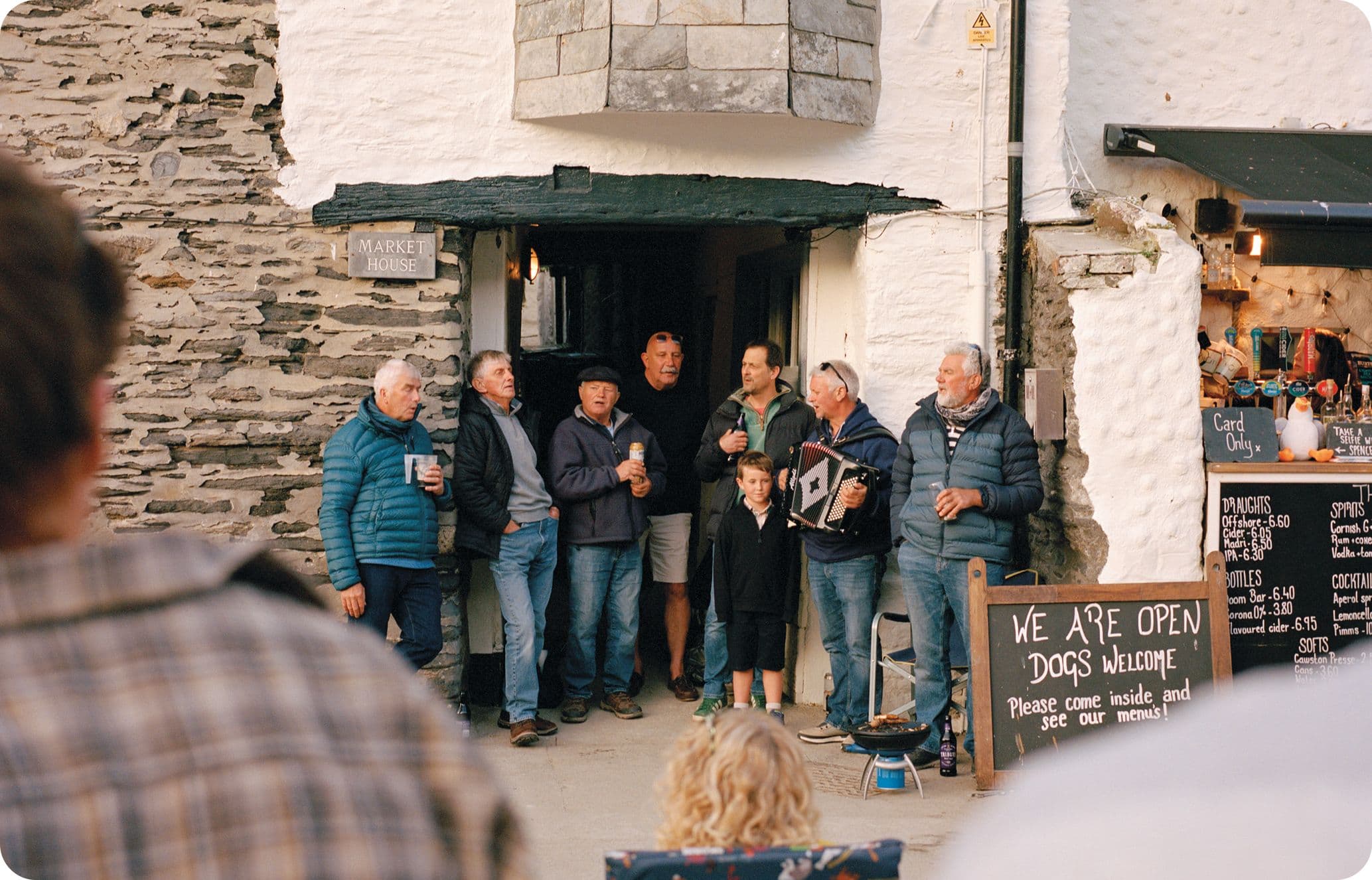 A group of people gather outside a rustic building, some holding drinks. A chalkboard sign invites dogs. A child stands among the group.