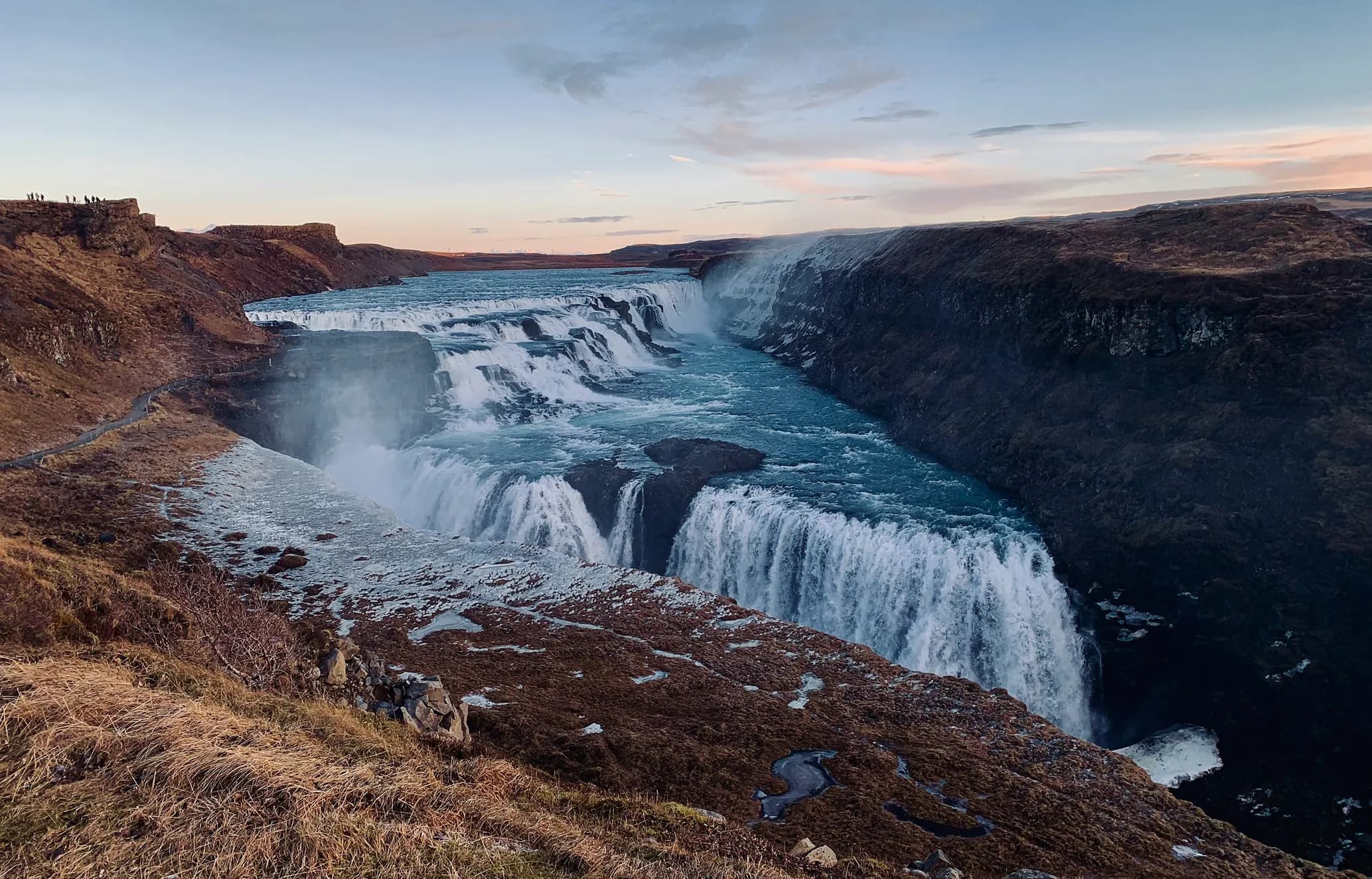 Gullfoss, Iceland: a powerful, multi-tiered blue waterfall plunges into a rugged canyon. Icy patches on brown land, people on a distant cliff.