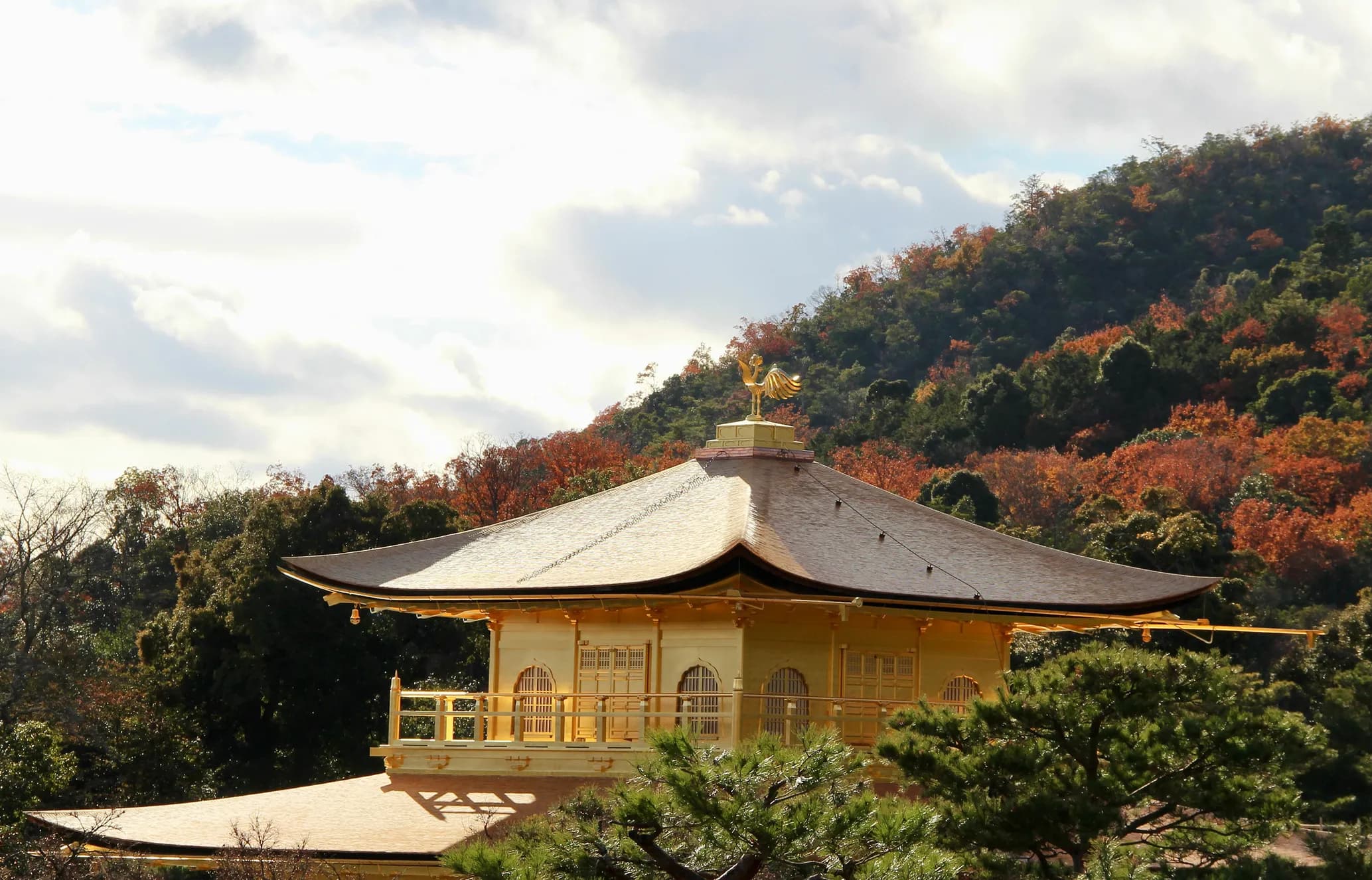 Golden temple roof with a phoenix statue, surrounded by autumn trees and hills under a cloudy sky.