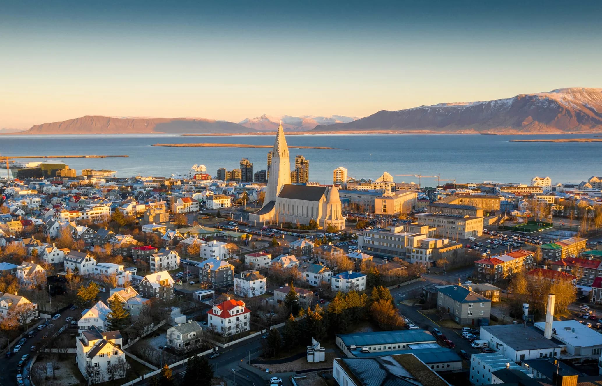 Aerial view of Reykjavik with the Hallgrímskirkja church, colorful houses, a bay, and snow-capped mountains under a golden sunset.