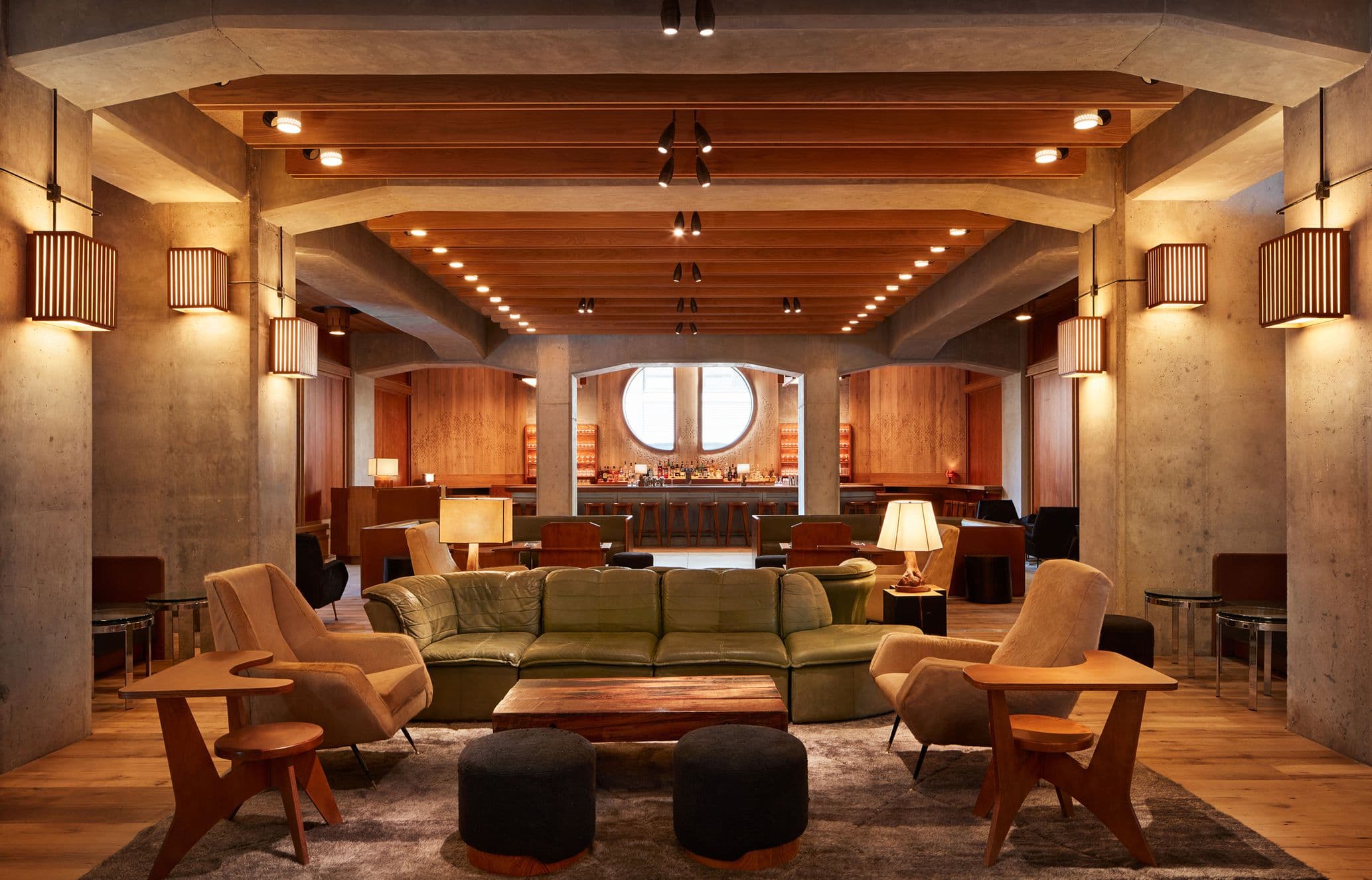 Cozy lounge with a green sofa, wooden chairs, a central coffee table, and warm lighting under wooden beam ceiling, in a modern, elegant space.