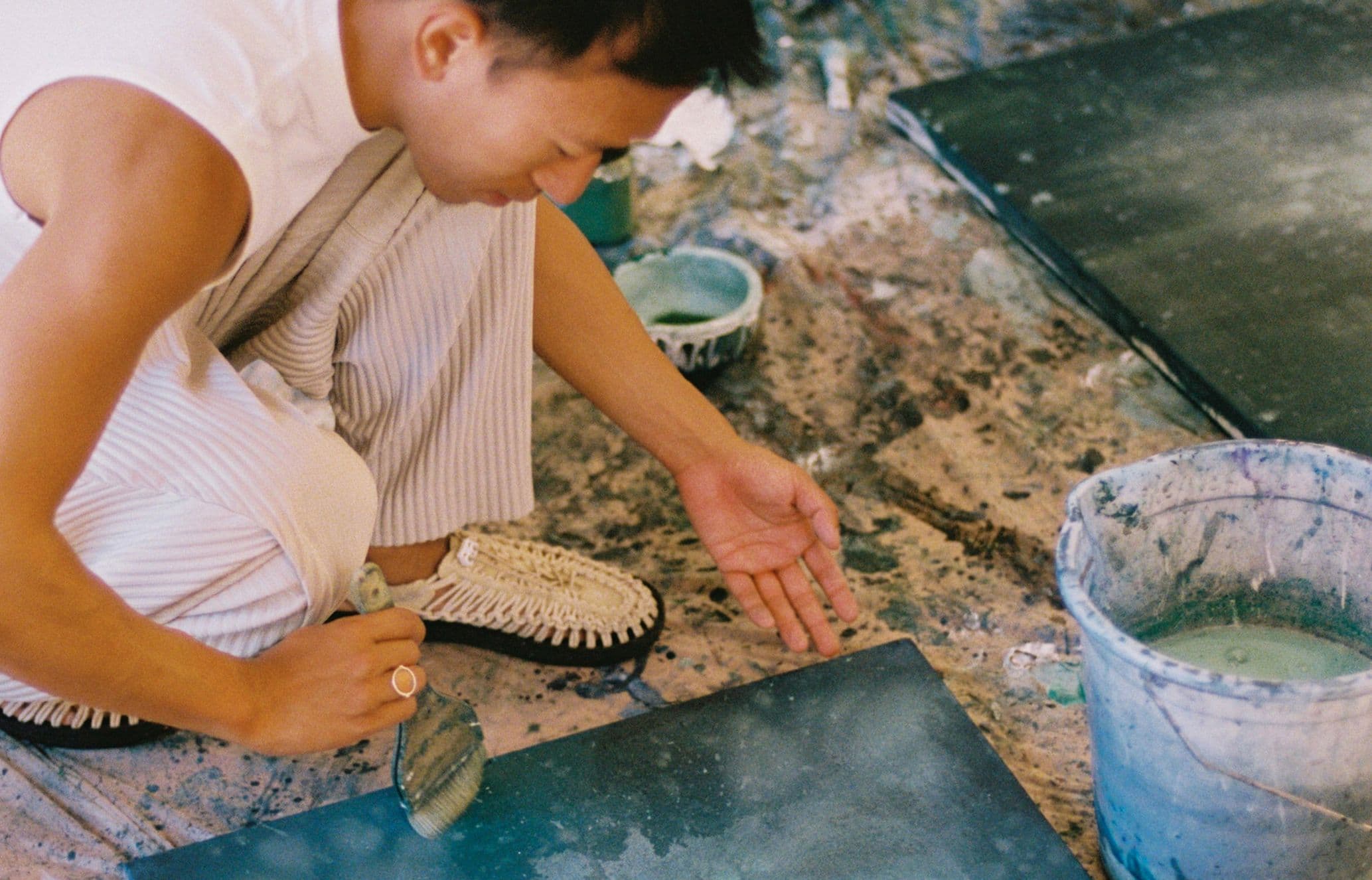 A person in white clothing crouches on a paint-splattered floor, working with green paint on a canvas using a trowel.