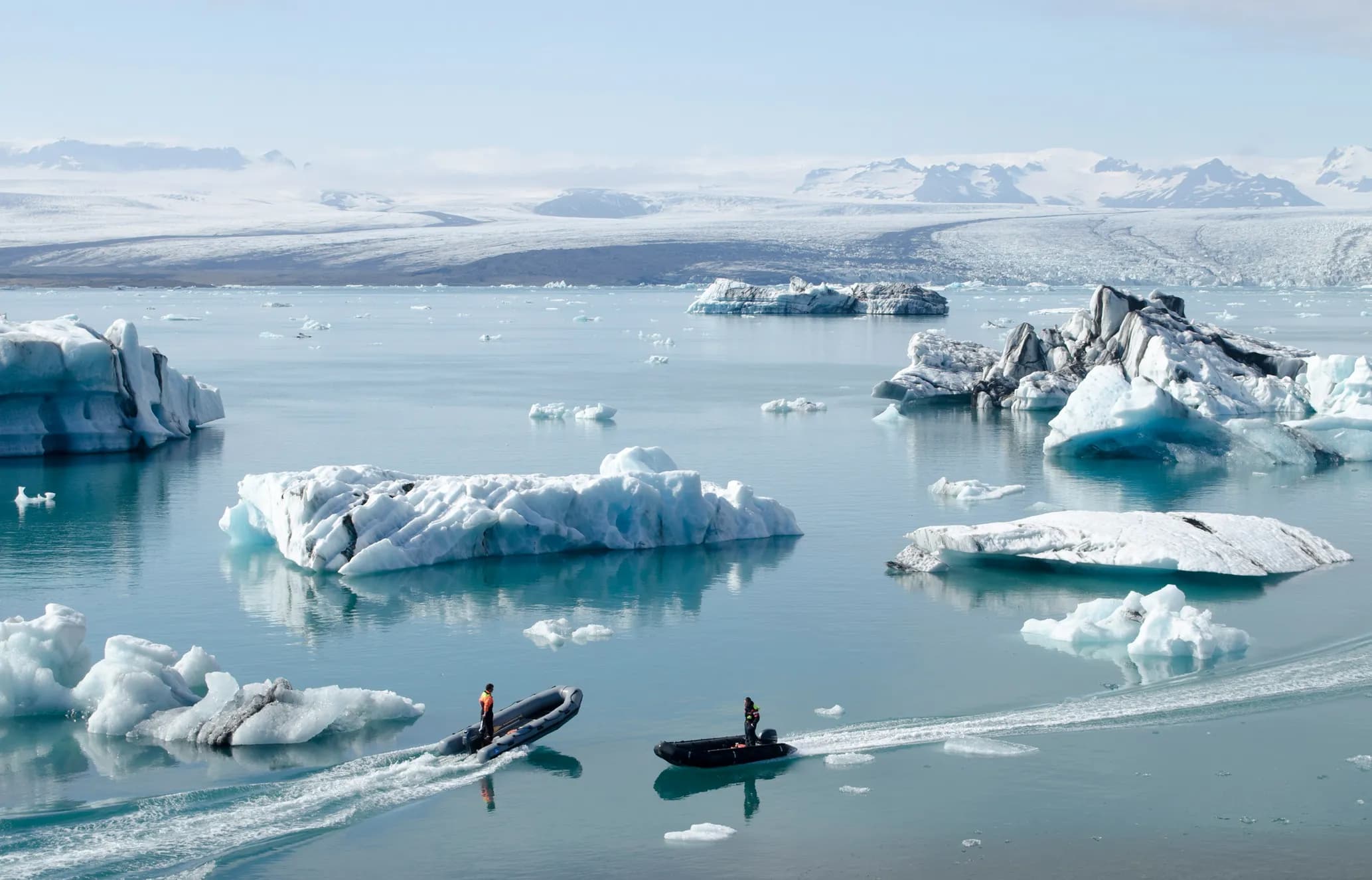 Two small boats speed through a glacial lagoon filled with numerous icebergs, some with blue hues, against a backdrop of snow-covered mountains.