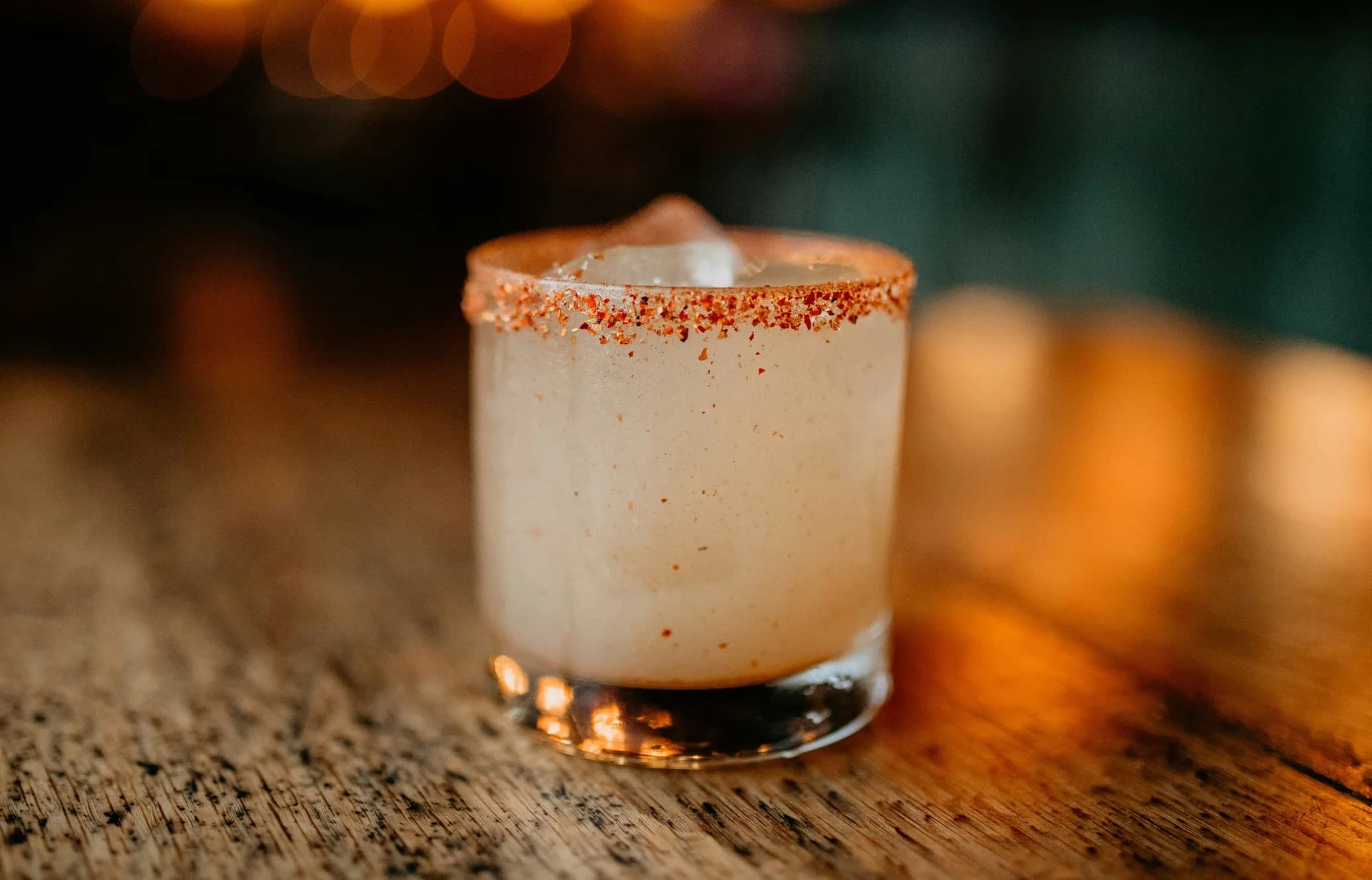 A cocktail in a glass with a chili-rimmed edge, filled with a frothy beverage, sits on a wooden table with warm lighting in the background.