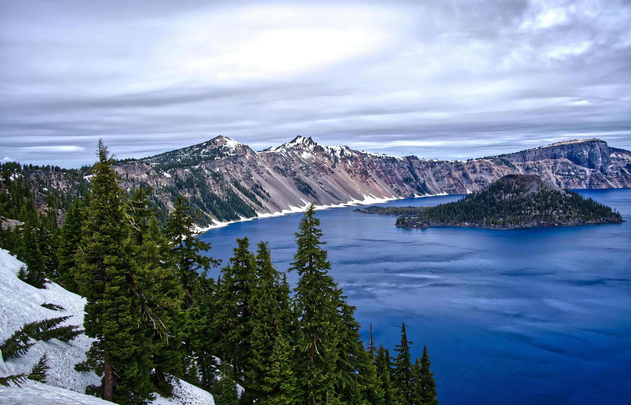 Snow-covered cliffs and pine trees overlook the deep blue waters of a lake, with a mountain range under a cloudy sky in the background.