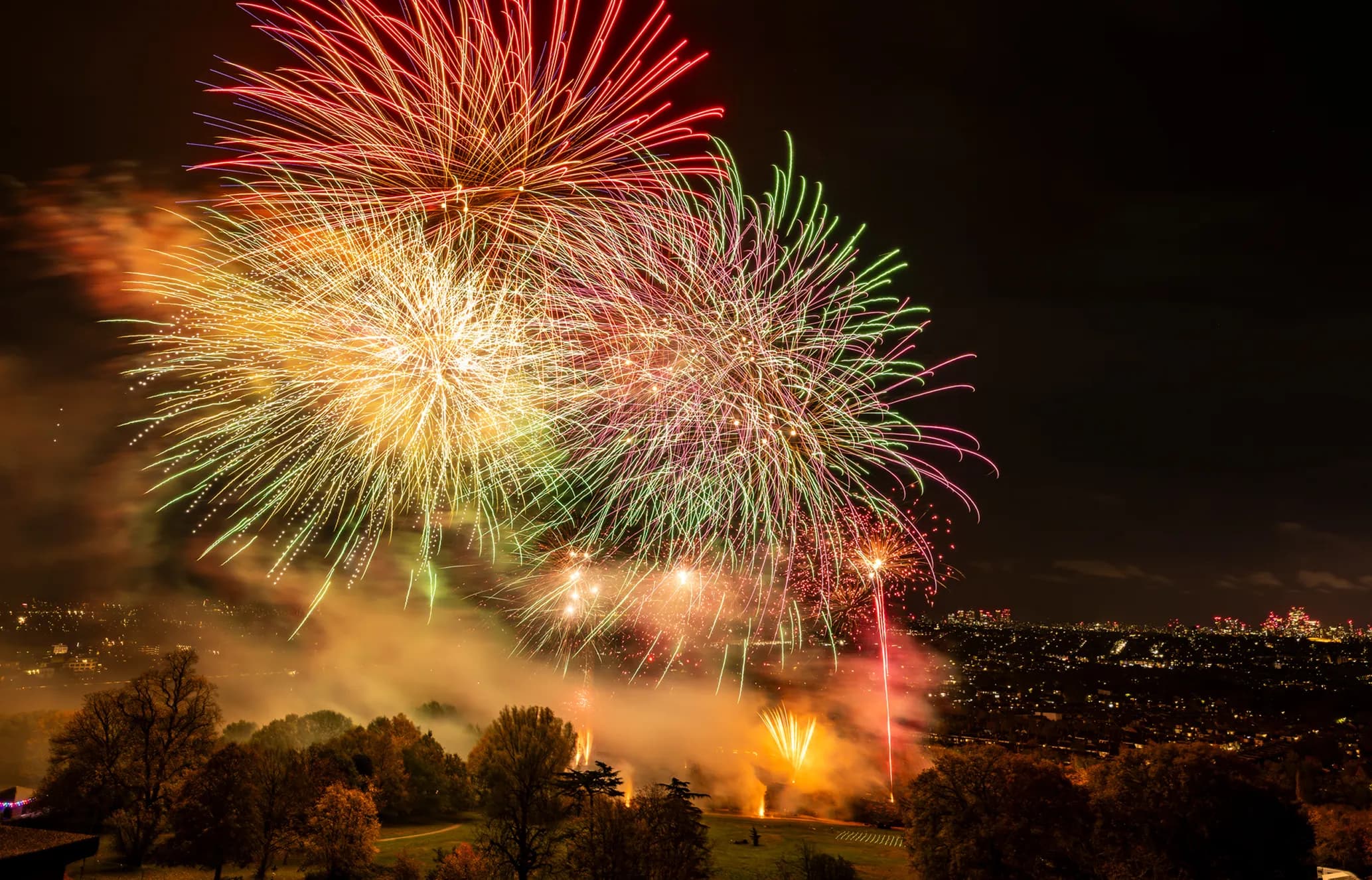 Colorful fireworks explode in the night sky over a cityscape, illuminating trees and a hill in the foreground.