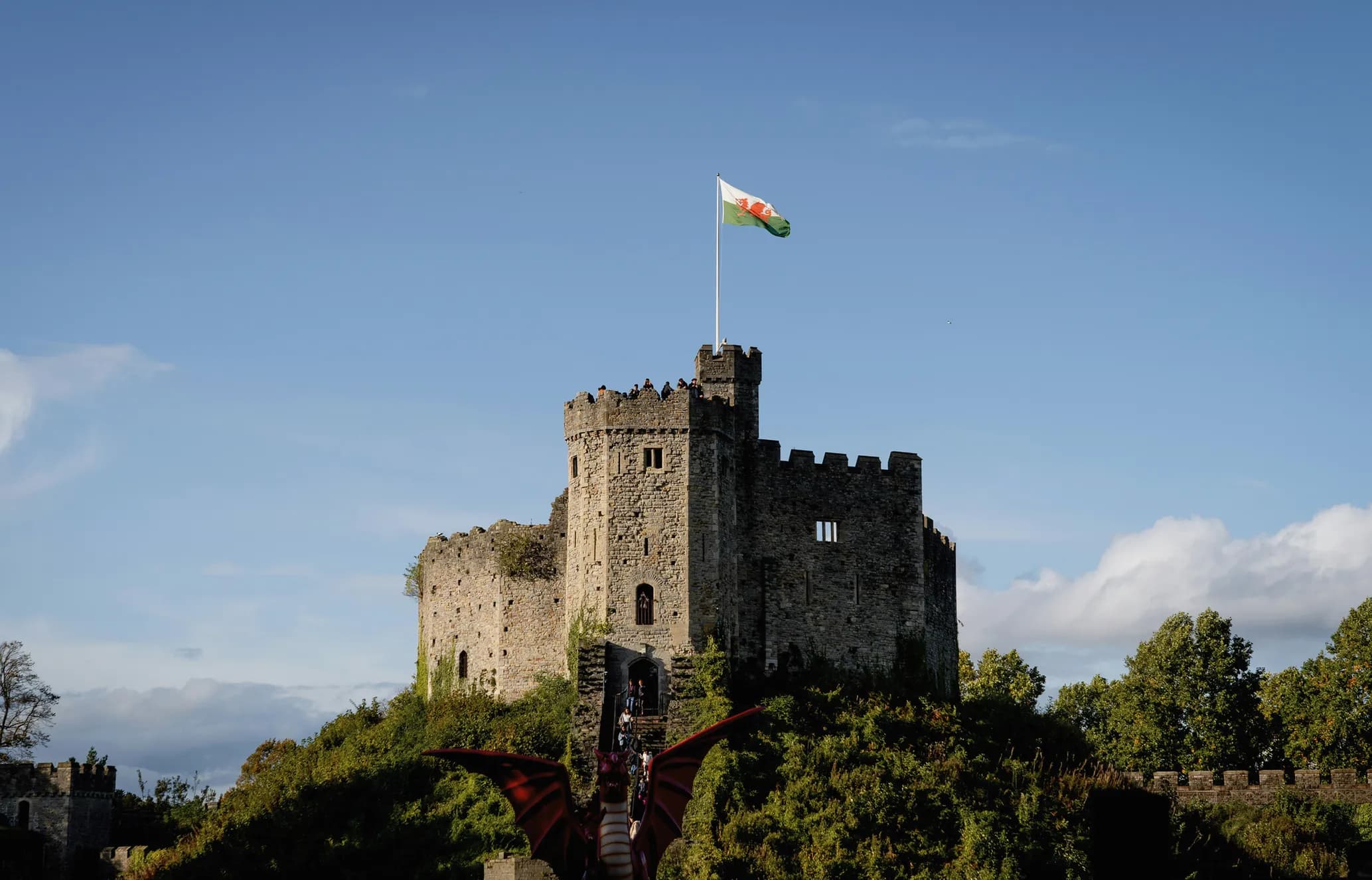 Stone castle with towers, a Welsh flag flying on top, surrounded by greenery; dragon wings visible in the foreground under blue sky.